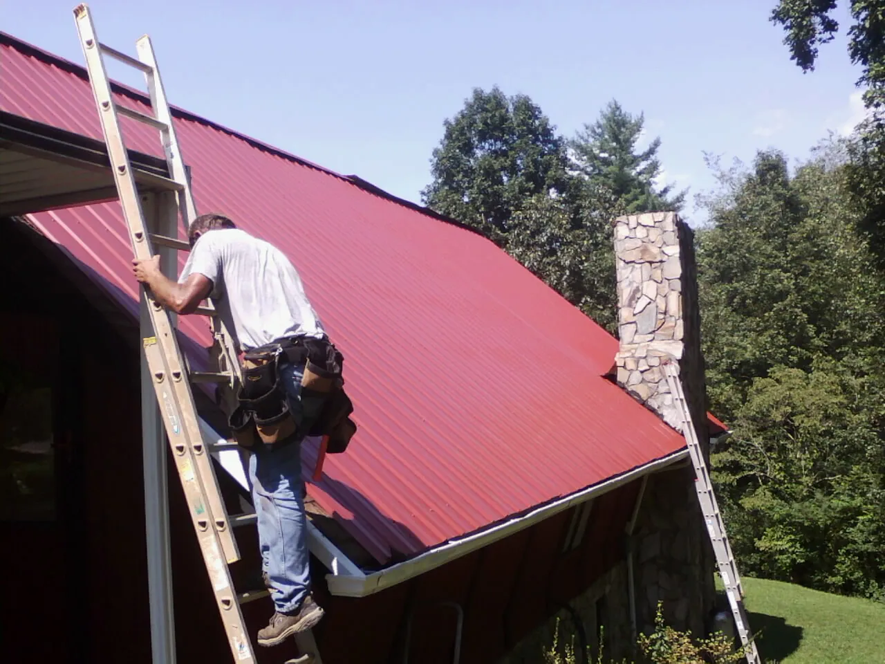 Roofer on a ladder beside a red metal roof and stone chimney — Soffit & Fascia Repair by Roofing Brothers in Belcamp